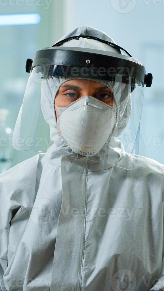 Tired microbiologist in coverall sitting in laboratory looking at camera. Team of scientits examining virus evolution using high tech and chemistry tools for scientific research, vaccine development. photo