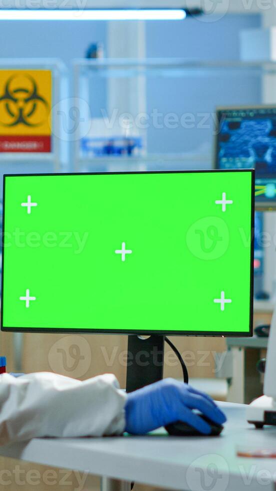 Scientist man wearing protection suit typing on computer with green mockup in modern equipped lab. Team of microbiologists doing vaccine research writing on device with chroma key, isolated display. photo