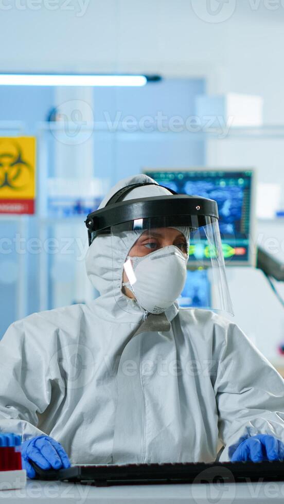 Scientist in coverall examining blood test tubes at laboratory DNA testing analysis writing at pc. Doctor working with various bacteria, tissue, pharmaceutical research for antibiotics against covid19 photo