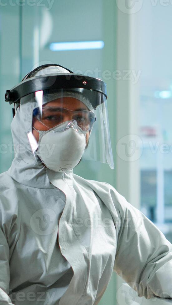 Tired laboratory doctor with overall suit working at computer and looking at camera. Scientist examining virus evolution using high tech, chemistry tools for scientific research, vaccine development photo