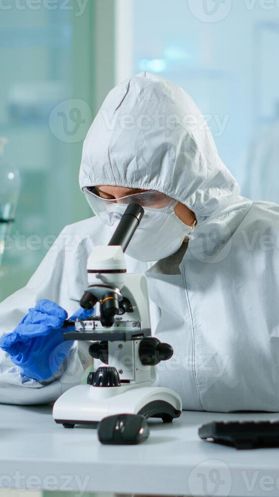 Biologist doctor in coverall checking DNA sample on microscope typing on pc in equipped lab. Examining vaccine evolution in medical lab using high tech and chemistry tools for scientific research photo