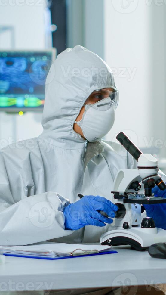 Medical research scientist conducting DNA experiments under microscope wearing protection suit and writing informations on clipboard. Chemist using high tech for vaccine development against covid19 photo