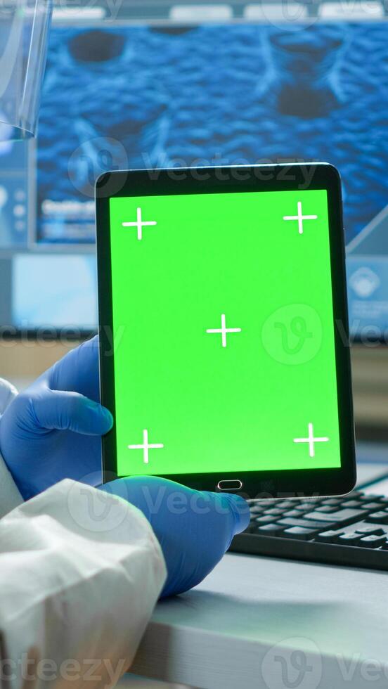 Chemist man wearing protection suit holding tablet with green mockup in modern equipped lab. Team of microbiologists doing vaccine research writing on device with chroma key, isolated display. photo