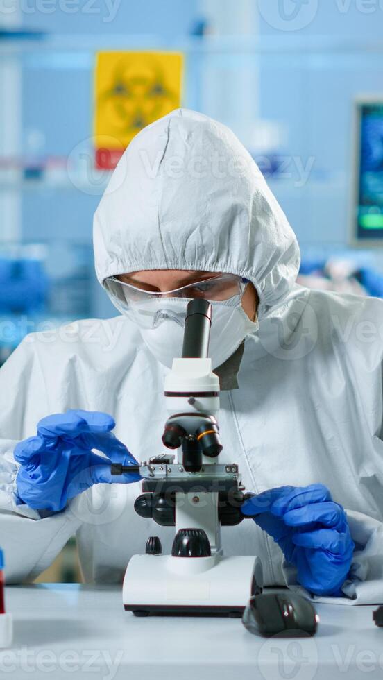 Scientist in ppe suit making adjustments and looking through laboratory microscope, typing on pc. Chemist in coverall working with various bacteria, tissue blood samples for antibiotics research photo