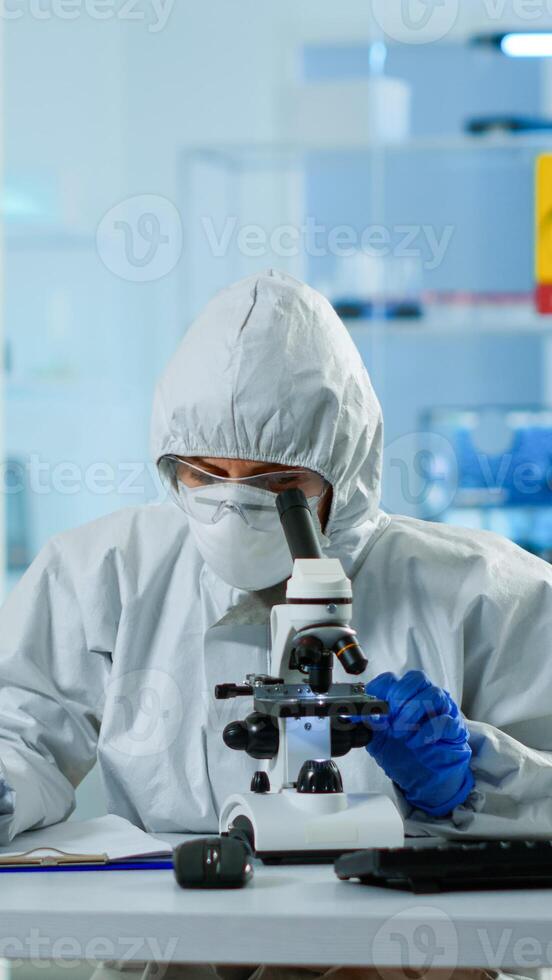 Biologist in ppe suit working on microscope and writing information on clipboard in equipped laboratory. Chemist examining vaccine evolution using high tech researching diagnosis against covid19 virus photo