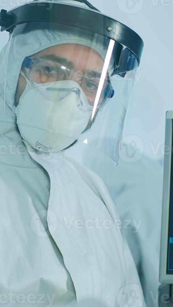 Microbiologist in ppe suit standing in laboratory looking at camera behind the glass wall in equipped lab. Doctor examining virus evolution using high tech and chemistry tools for vaccine development photo