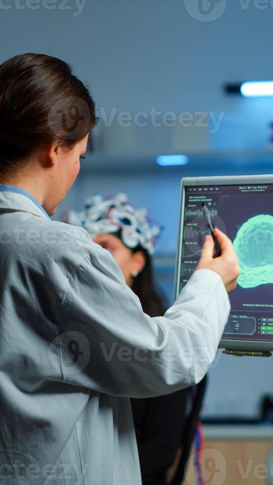 Researcher looking at monitor analysing brain scan while coworker discussing with patient in background about side effects, mind functions, nervous system, tomography scan working in laboratory photo