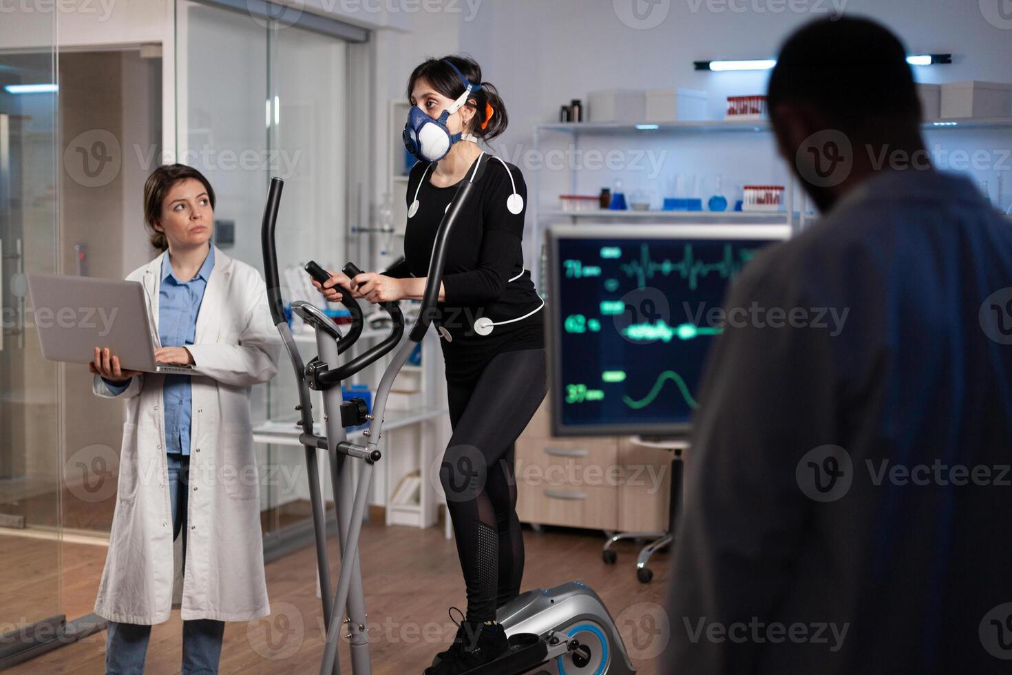 Specialist fitness researcher examining endurance controling EGK of athlete woman with mask running on bycle trainer. Physician man analyzing heart rate in medical modern laboratory. Medicine service photo