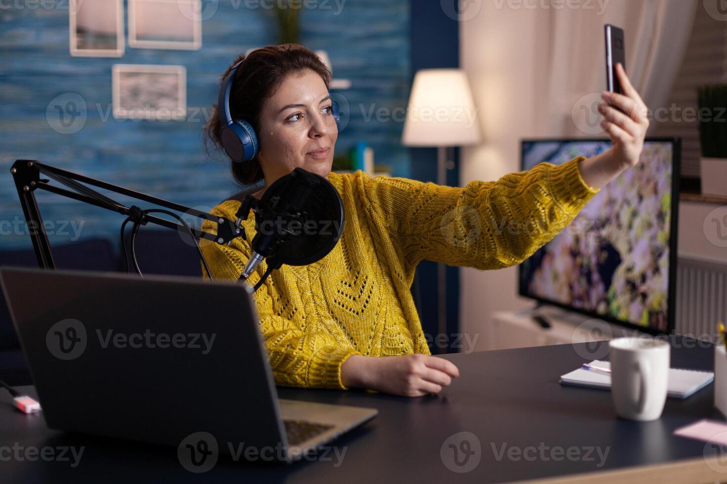 Social media influencer taking selfie while recording entertainment podcast using vlog equipment in living room studio. Woman vlogger talking into podcasting microphone. On-air show photo