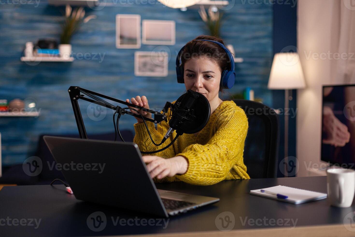 Portrait of social media influencer wearing studio headphones talking with online listeners using professional microphone during livestream at home in living room. Woman host recording production photo
