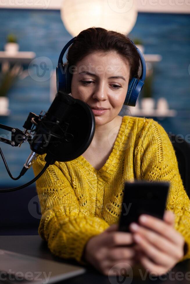 Portrait of social media influencer with headphones holding smartphone reading questions answering using production microphone sitting at desk in podcasting studio at home. On-air show photo