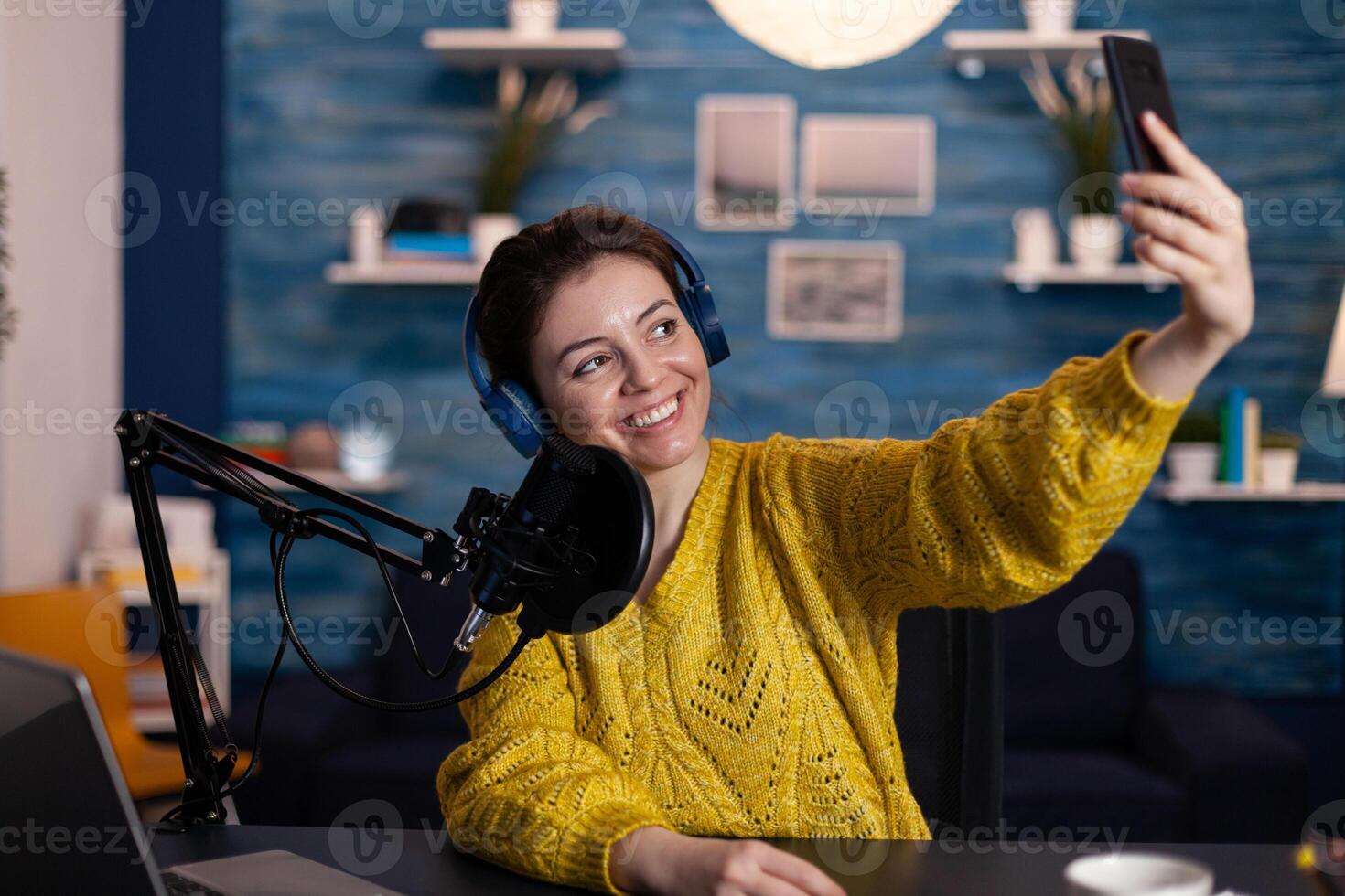 Social media blogger with headphones taking selfie while recording entertainment podcast using vlog equipment in living room studio. Woman influencer talking into podcasting microphone. On-air show photo