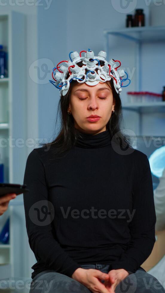 Stressed patient sitting on neurological chair with eeg headset, researcher examining health status writing on tablet. Medical scientist looking through microscope in background developing treatment photo