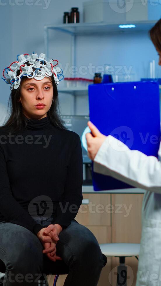 Neuroscience doctor holding clipboard showing treatment against brain disease to patient with eeg headset. Woman sitting in neurological scientific laboratory treating nervous system dysfunctions photo