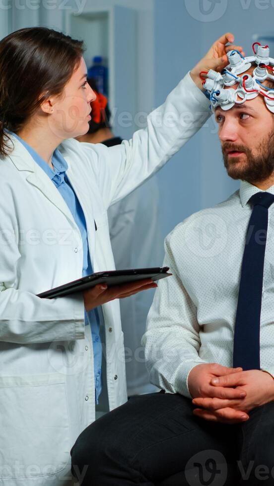 Man sitting on neurological chair with brainwave scanning headset, researcher doctor tracking evolution of patient's brain using tablet, adjusting eeg device. Scientist examining nervous system. photo