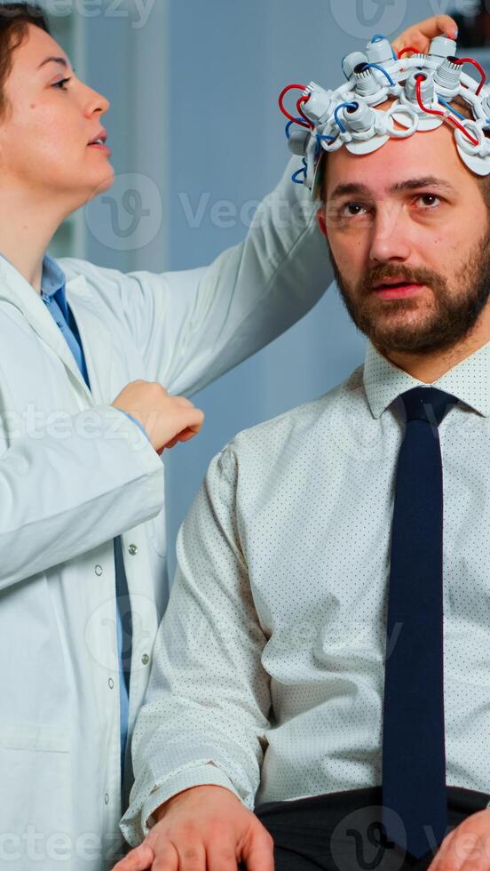 Patient with brainwave scanning headset sitting in brain study laboratory while neurologist scientist adjusting high tech device. Man visiting professional doctor treating nervous system dysfunctions photo