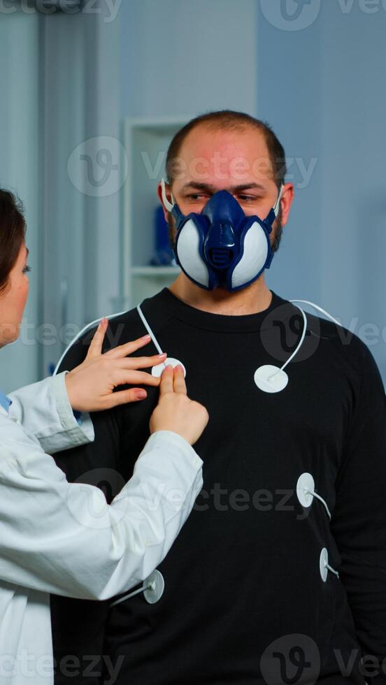 Woman researcher preparing patient for endurance test using electrodes and professional body equipment. Team of doctors monitoring health of man with special mask, ekg scan runs on computer screen. photo