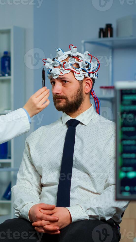 Man sitting on neurological chair with brainwave scanning headset while researcher examining health status writing on clipboard. Doctors working in brain study lab with monitors showing EEG reading. photo