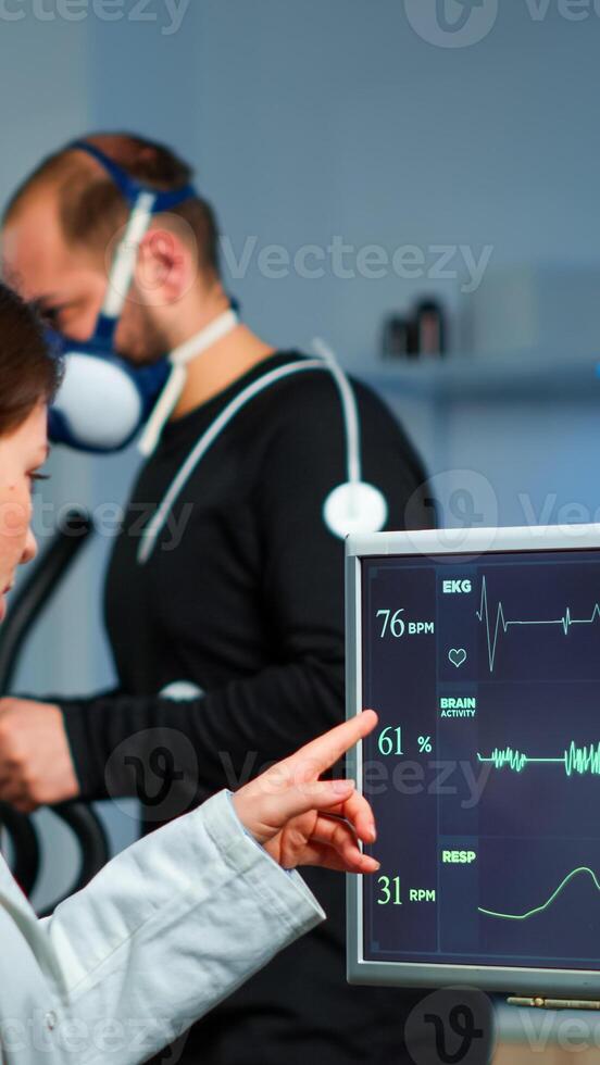 Muscular athlete with mask running on cross trainer in medical laboratory for monitoring his performance while ekg scan runs on computer screen and team of doctor researcher examining results photo