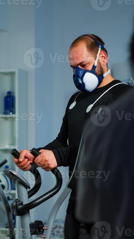 Patient making electrocardiogram during physical exercise in sports science lab, monitoring endurance and heart rate, computer showing EKG data on display. Man running with mask and medical electrodes photo