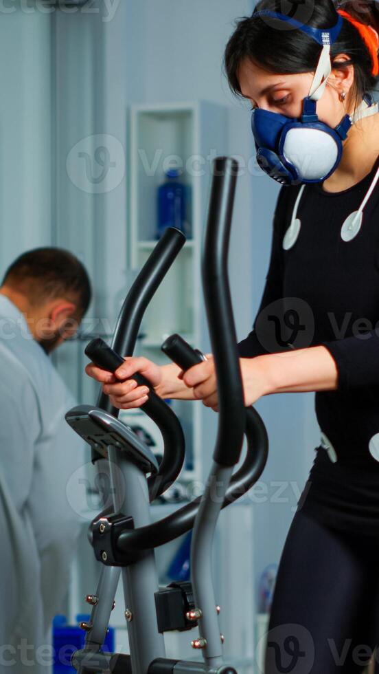 Woman athlete with mask running on cross trainer in sports science lab measuring performance and oxygen consumption during Vo2max test. Medical researcher looking at EKG scan monitoring heart rate photo