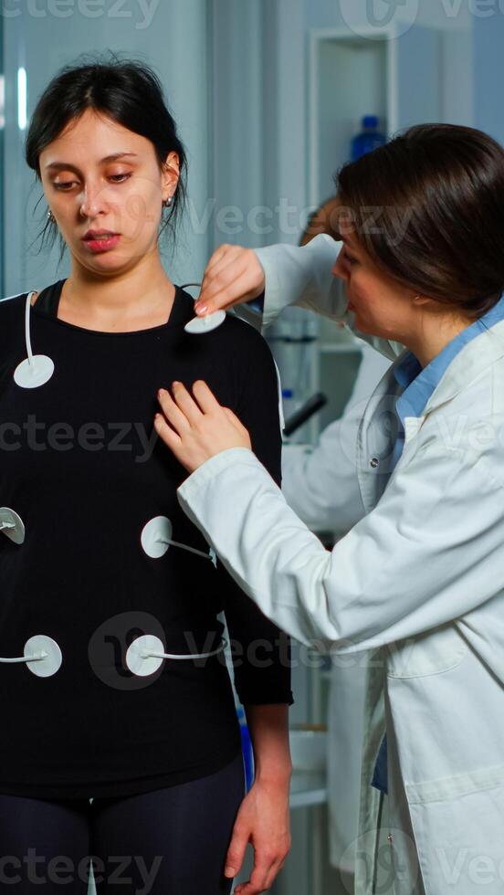 Scientist researcher preparing woman patient for endurance test attaching electrodes on professional body equipment. Team of doctors monitoring health of patinet, vo2, ekg scan runs on computer screen. photo
