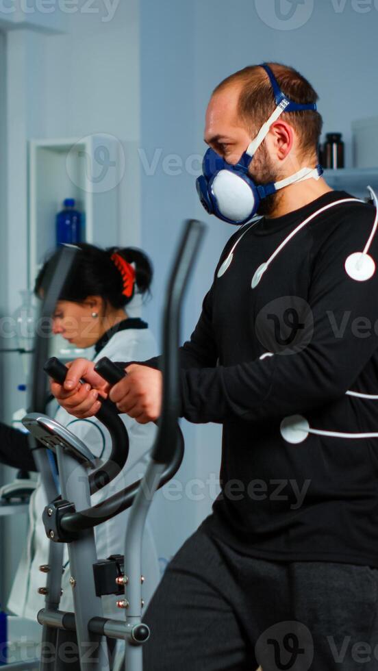 Sportsman running on cross trainer in lab facility for sports performance, medical researcher monitoring endurance and heart rate. Monitor showing EEG, EKG reading of athlete running on cross trainer photo