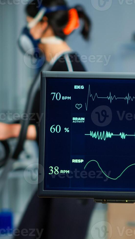 Medical researcher examining EKG image showing on monitor while patinet with mask running on cross trainer testing heart rate using electrodes.Doctor monitoring physical endurance in science sport lab photo