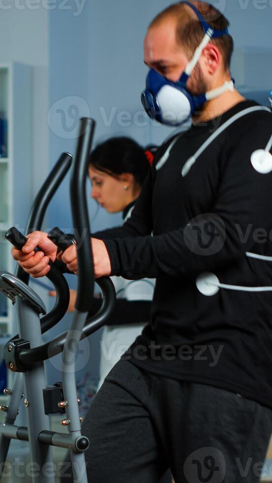 Science sport doctor asking patient about his health while sportsman running on cross trainer with mask and electrodes attached on body monitoring vo2. Woman researcher measuring endurance of patient. photo