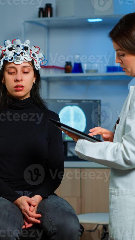 Researcher neurologist doctor asking patient's symptoms taking notes on tablet, adjusting high tech eeg headset. Doctor researcher controlling EEG headset analyzing brain functions and health status photo