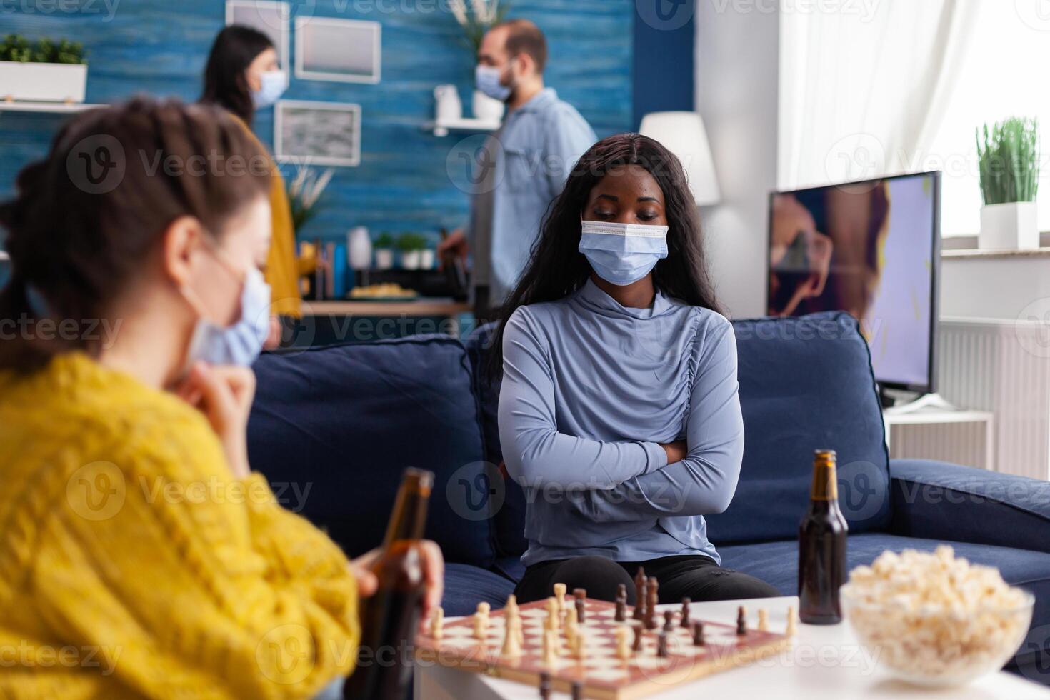 African woman wearing face mask playing chess drinking beer, keeping social distancing preventing spread of covid virus. Diverse people having fun in home living room playing board games photo