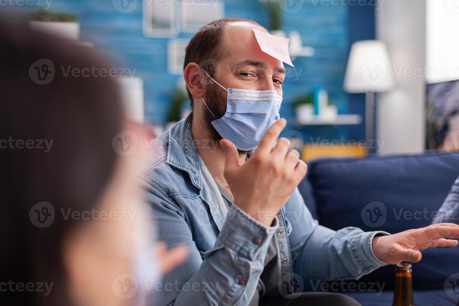 Thoughtful multiethnic friends with sticky notes on foreheads playing name game miming gesturing wearing face mask keeping social distancing preventing covid19 spread. People enjoying bottles of beer photo