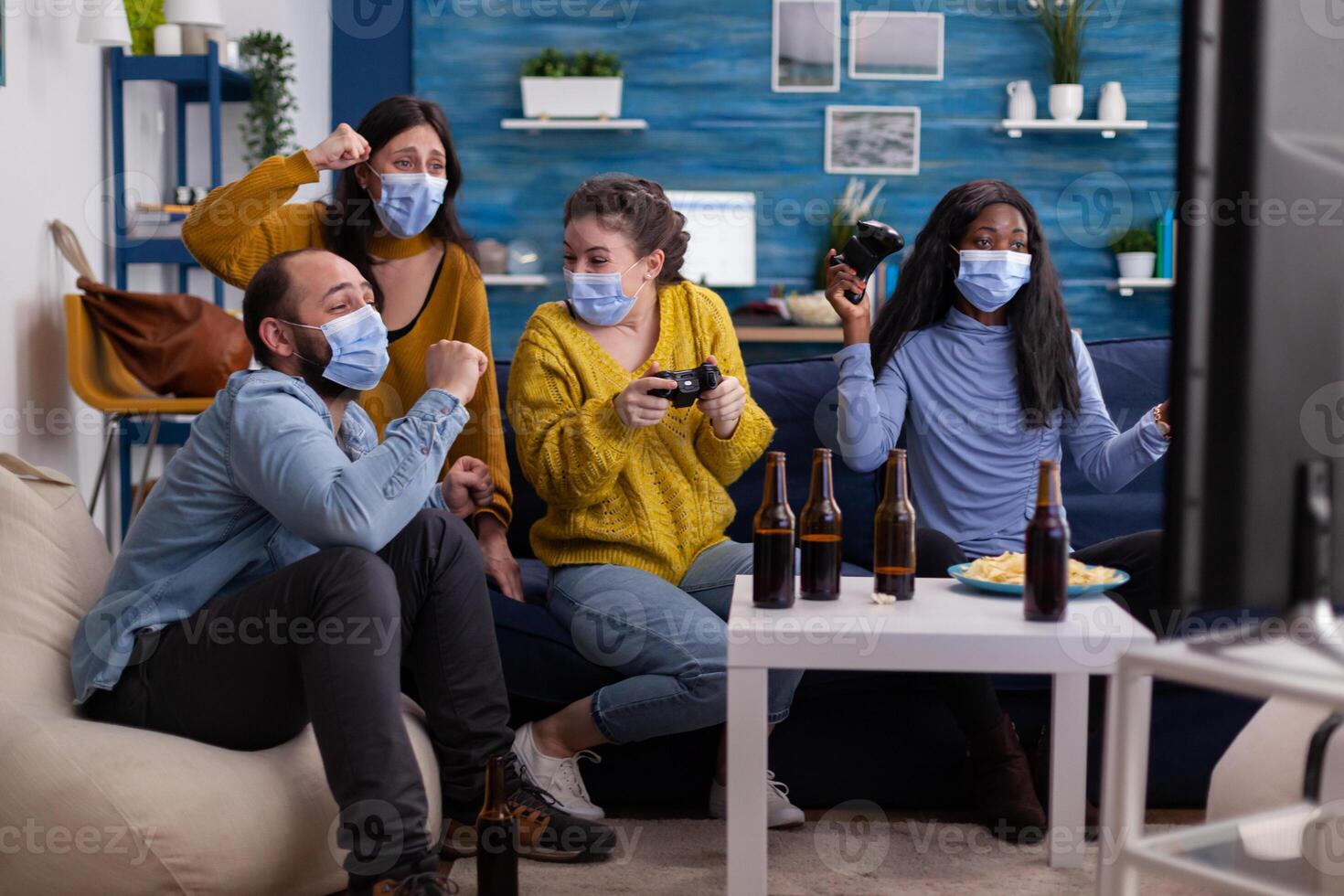 Group of multiethnic friends celebrating victory while playing games with joypad in living room keeping social distancing wearing face mask during global pandemic. People enjoying time together. photo