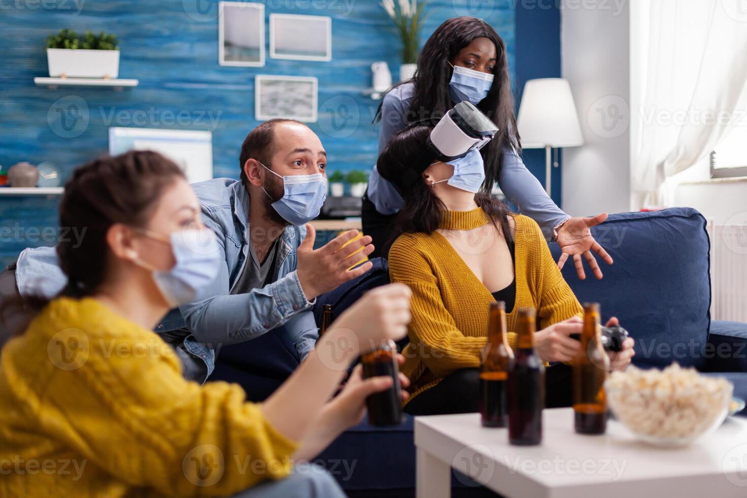 Man with face mask cheering up for friends playing games with vr headset in home living room keeping social distancing in time of global pandemic with covid. Multi ethnic people having fun. photo