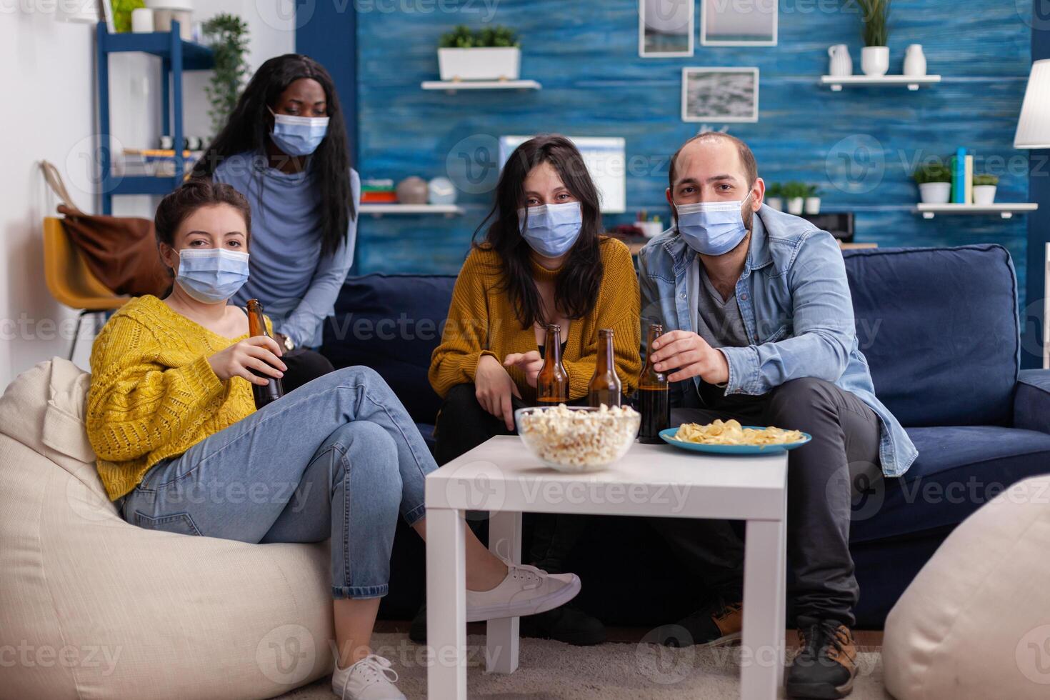 Pov of african woman and friends looking at tv holding remote control wearing face mask because of coronavirus pandemic outbreak drinking beer, relaxing, sitting on sofa in living room. photo