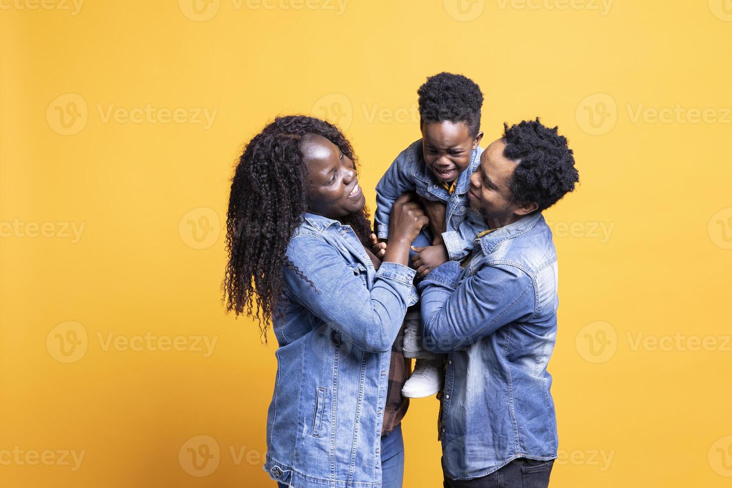 Happy african american partners lifting their little child on camera, posing on yellow background. Cheerful proud parents bonding with their adorable small kid, quality leisure time. photo