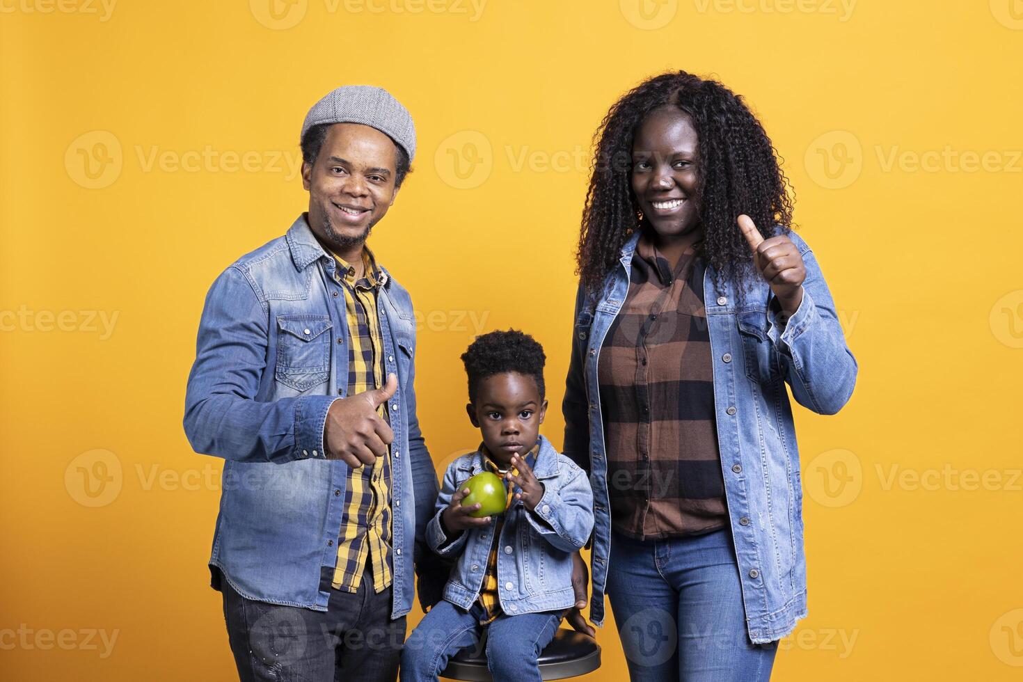 African american parents doing thumbs up symbol in studio, posing next to their cute little boy on the chair. Pleased grateful mother and father giving like gesture, agreeing on yellow background. photo