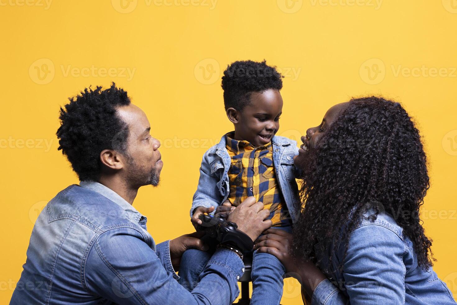 Happy small toddler playing around with a controller and sitting next to his parents, posing together in front of the camera. African american cheerful family of three against yellow background. photo
