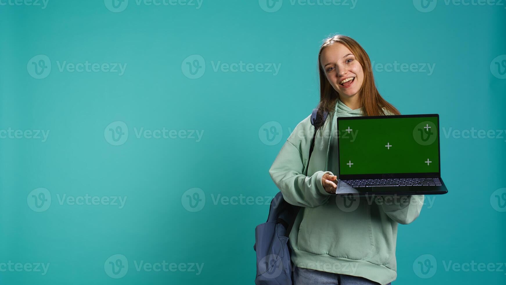 Smiling woman presenting laptop with green screen display, isolated over blue studio background. Cheerful person creating promotion with chroma key notebook device, camera B photo