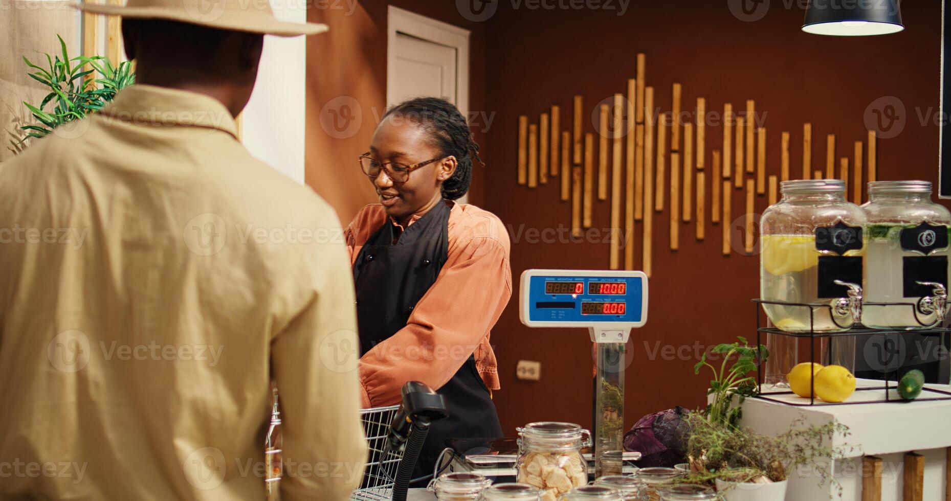 Woman scanning organic goods for client at checkout counter, selling locally grown pesticides free produce and pantry supplies. African american worker helping buyer to weight merchandise. Camera 2. photo
