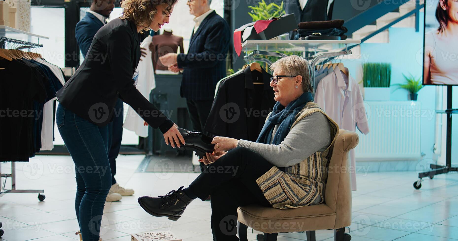 Retail worker presenting various pairs of shoes for senior client, assisting woman to find the perfect footwear in fashion showroom. Elderly customer trying on footgear before buying. Camera B. photo
