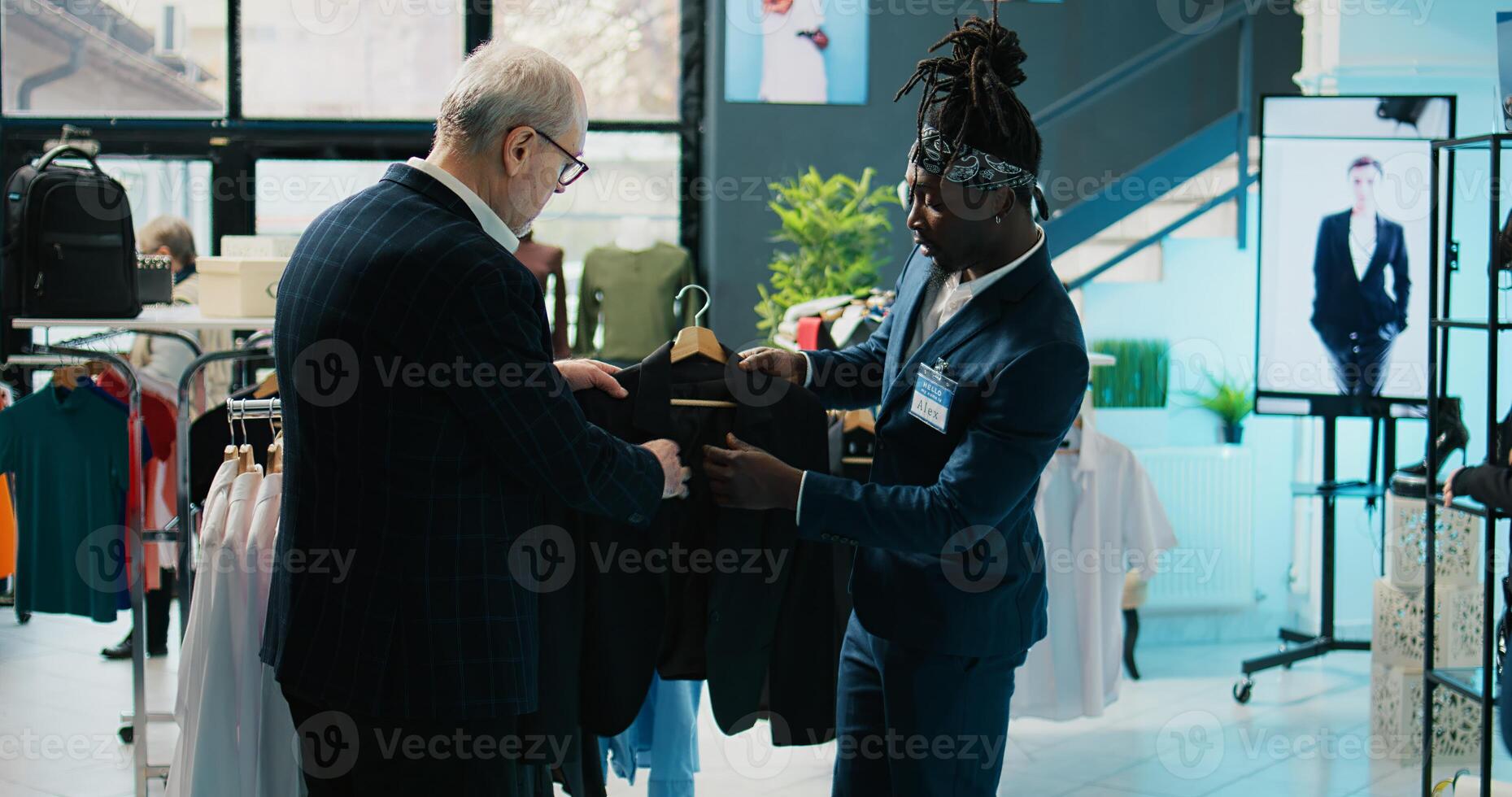African american store assistant helping regular client with a suit in shopping center, senior customer looking to buy formal clothes. Elderly man shopping for new collection at the mall. Camera B. photo