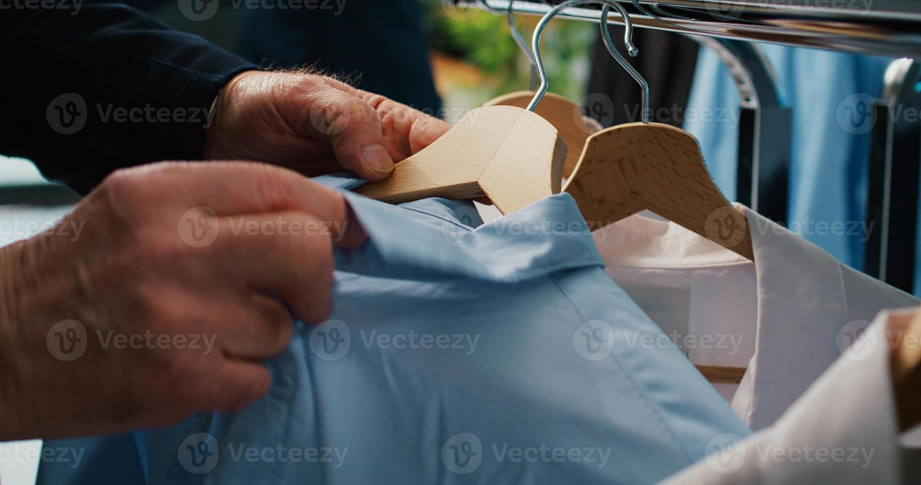 Elderly client checking all clothes placed on display at showroom, browsing through hangers to find items on discount. Senior man shopping for elegant shirts and suit jackets. Close up. Camera B. photo