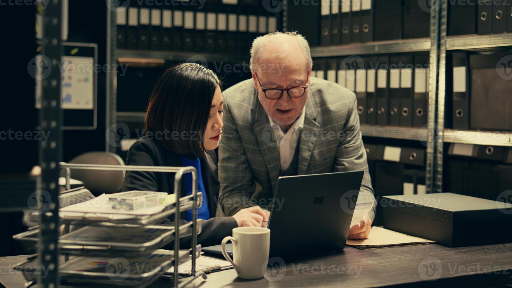 Senior inspector showing case details to his partner in archive room, studying forensic evidence and classified criminal records in a box. Detectives team work on federal investigation. Camera B. photo