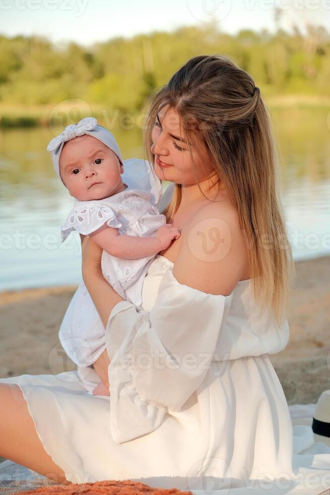 a beautiful girl in a white dress with a newborn baby is sitting on the beach, on the sand. photo