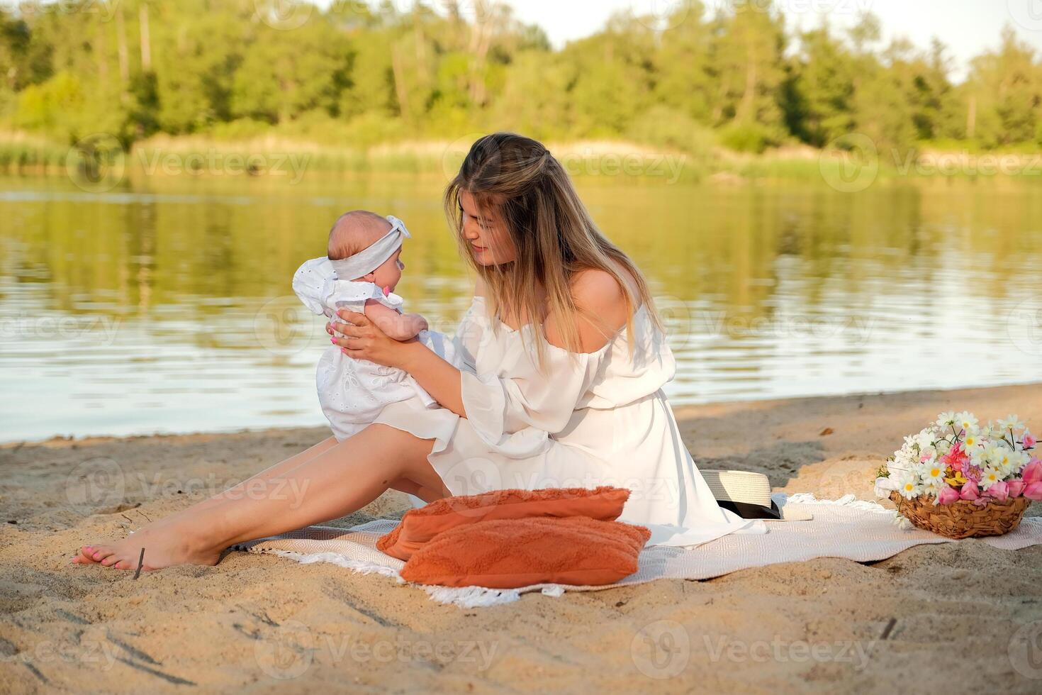 a beautiful girl in a white dress with a newborn baby is sitting on the beach, on the sand. photo