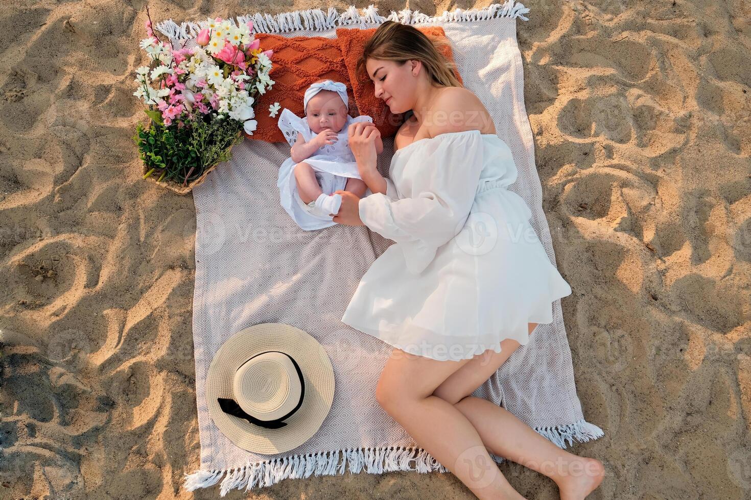 mom with a newborn baby lie on the beach. white dresses. flowers lie nearby. photo