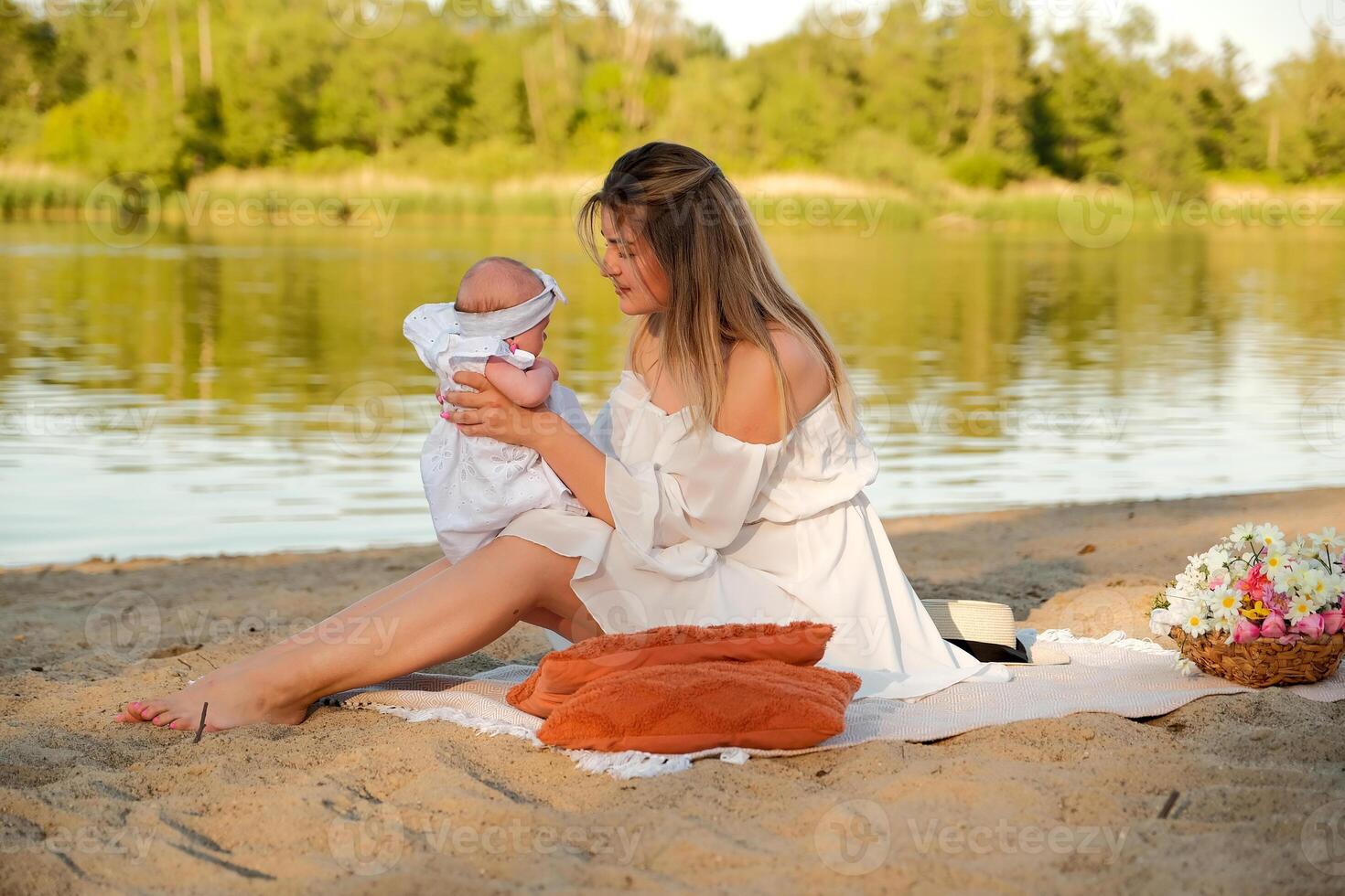 a beautiful girl in a white dress with a newborn baby is sitting on the beach, on the sand. photo