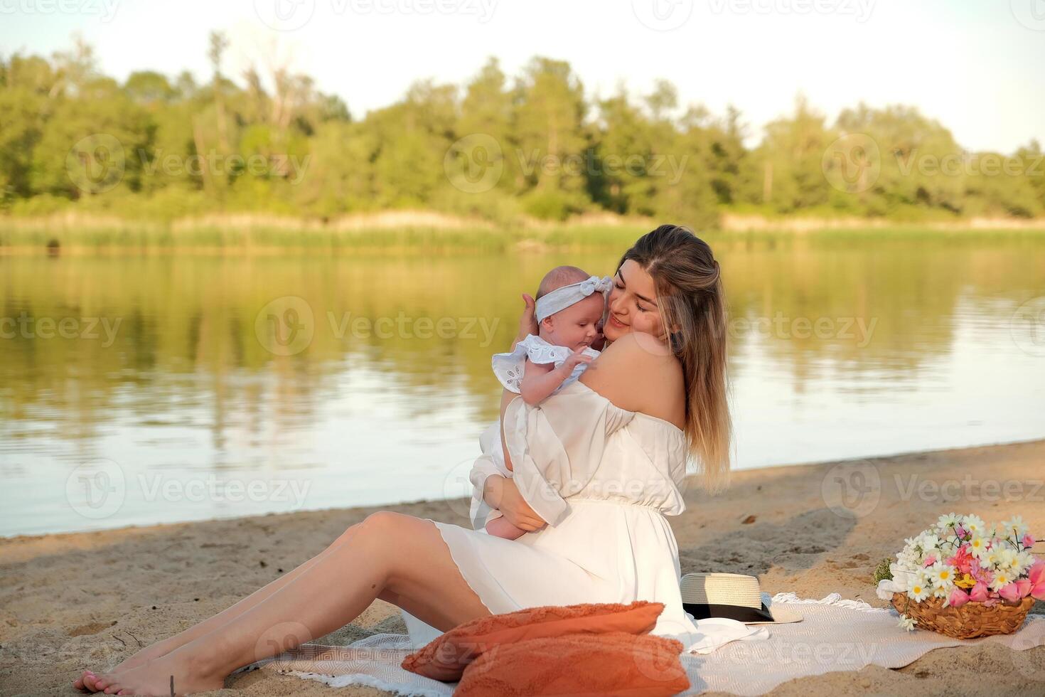 a beautiful girl in a white dress with a newborn baby is sitting on the beach, on the sand. photo
