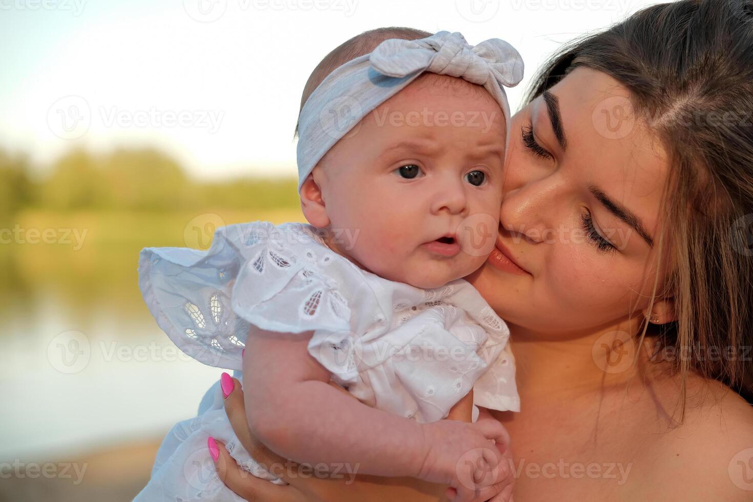a beautiful girl in a white dress with a newborn baby is sitting on the beach, on the sand. photo